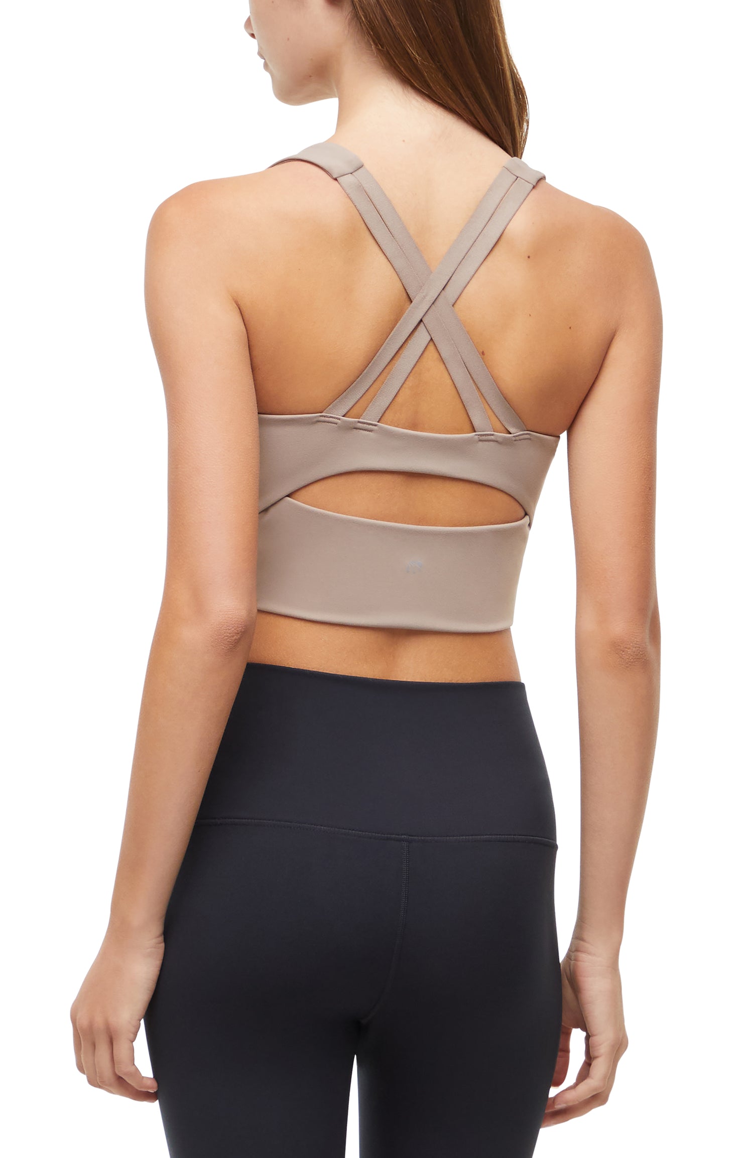 Back view of woman wearing a beige sports bra with criss-cross back design and black leggings on a white background. 