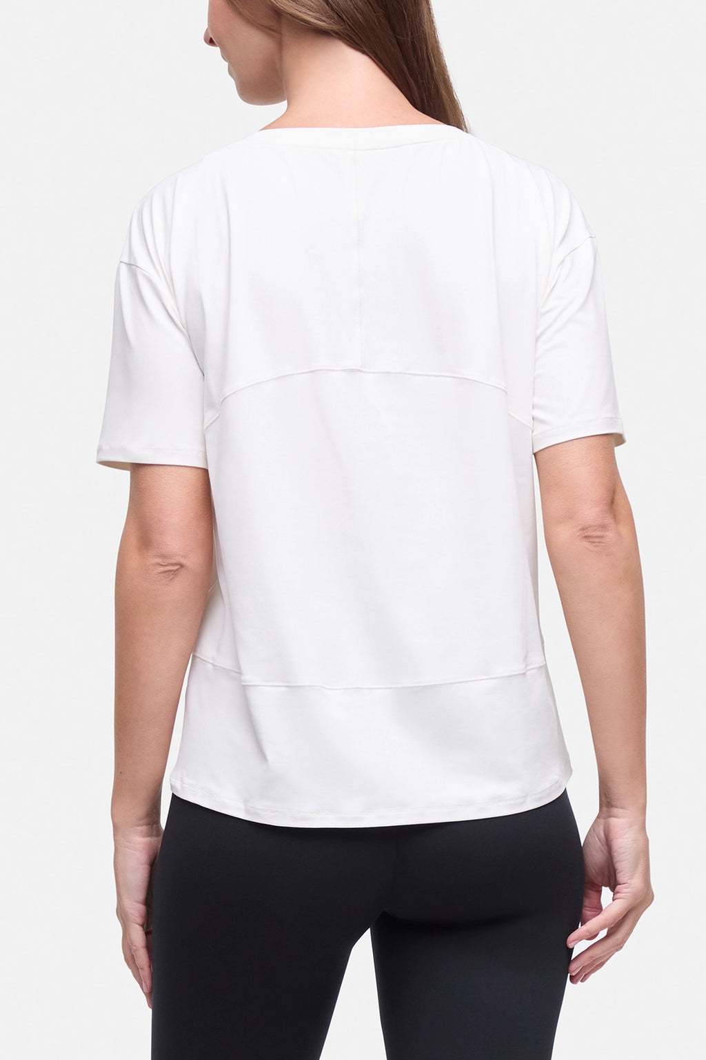 A woman shown from the back wearing a white short-sleeve athletic top with layered horizontal seam details and black leggings, against a light gray studio background. #color_sugar-swizzle