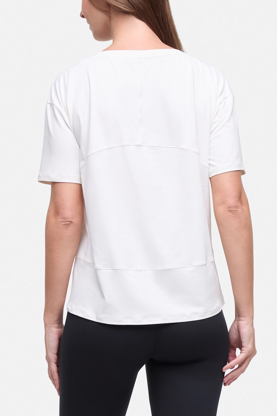 A woman shown from the back wearing a white short-sleeve athletic top with layered horizontal seam details and black leggings, against a light gray studio background. #color_sugar-swizzle