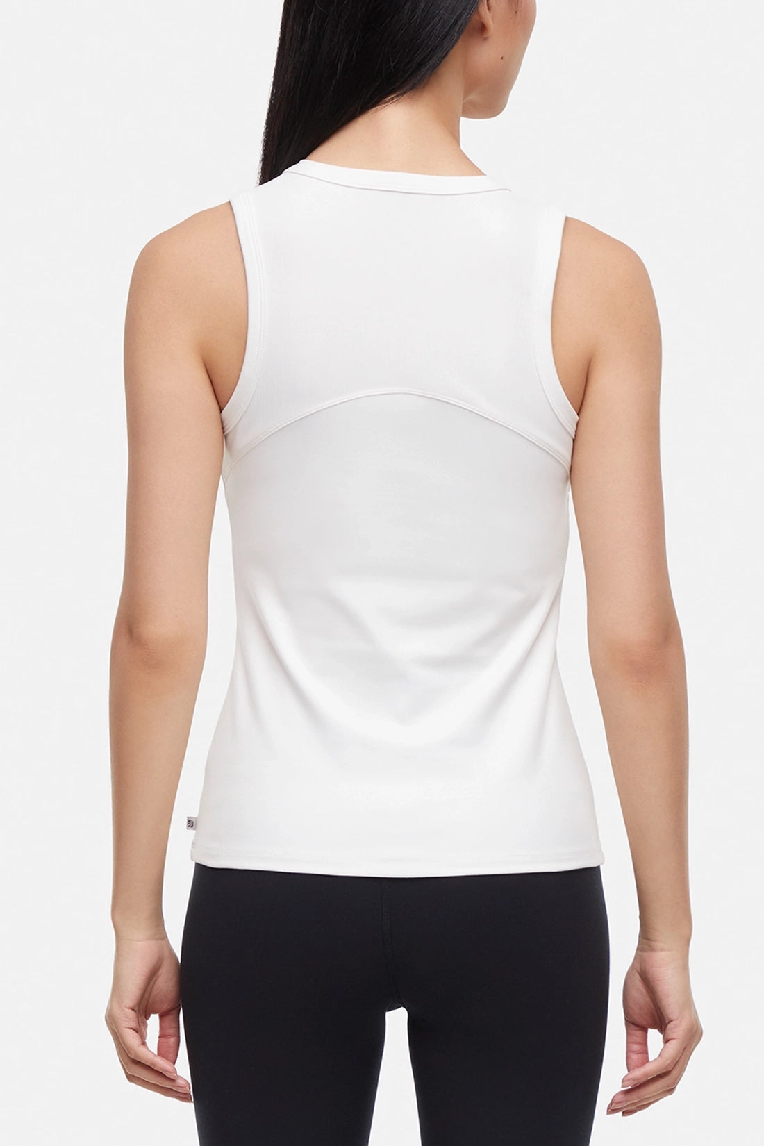 Back view of woman wearing a white ribbed-knit sleeveless tank top and black leggings on a light gray background.