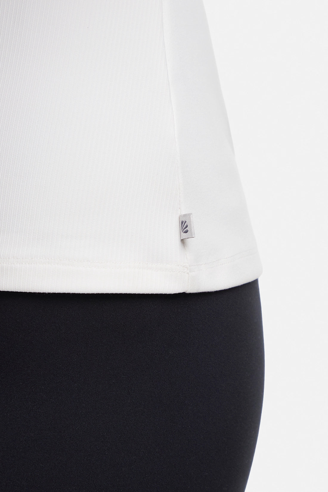 Close up view of woman wearing a white ribbed-knit sleeveless tank top logo tag and black leggings on a light gray background. #color_sugar-swizzle