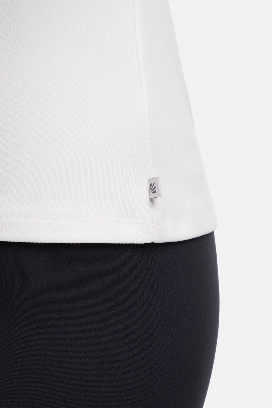 Close up view of woman wearing a white ribbed-knit sleeveless tank top logo tag and black leggings on a light gray background. #color_sugar-swizzle