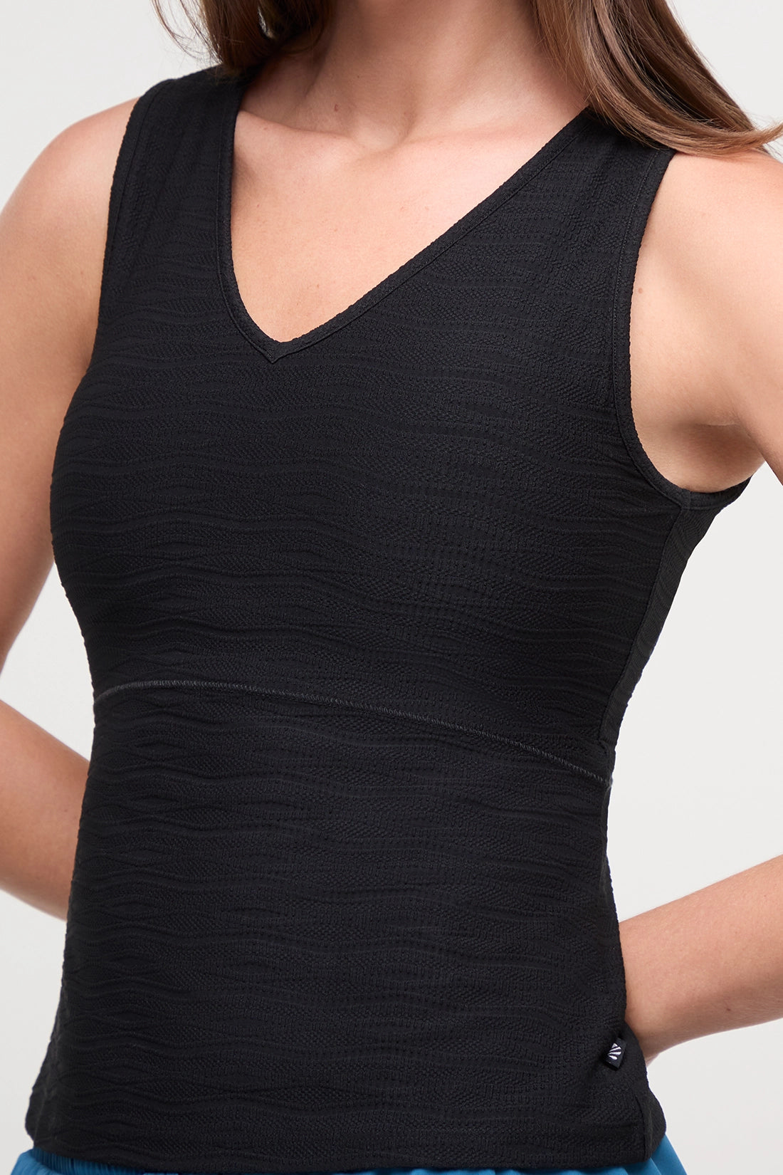 A woman wearing a black sleeveless V-neck athletic tank top and bright blue athletic shorts, shown from the shoulders to the upper thighs against a light gray studio background. 