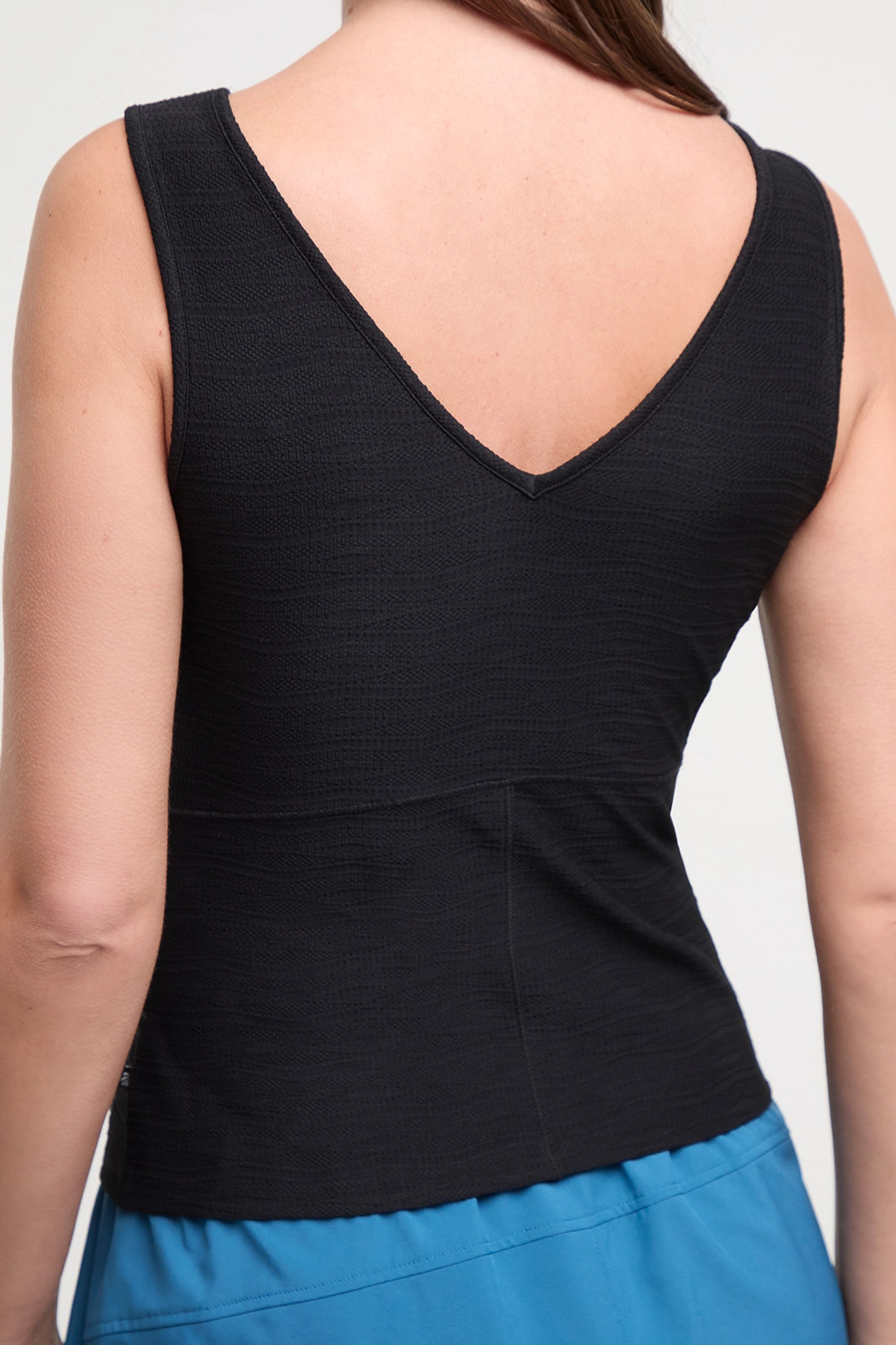 Back view of woman wearing a black sleeveless V-neck athletic tank top and bright blue athletic shorts, shown from the shoulders to the upper thighs against a light gray studio background. 