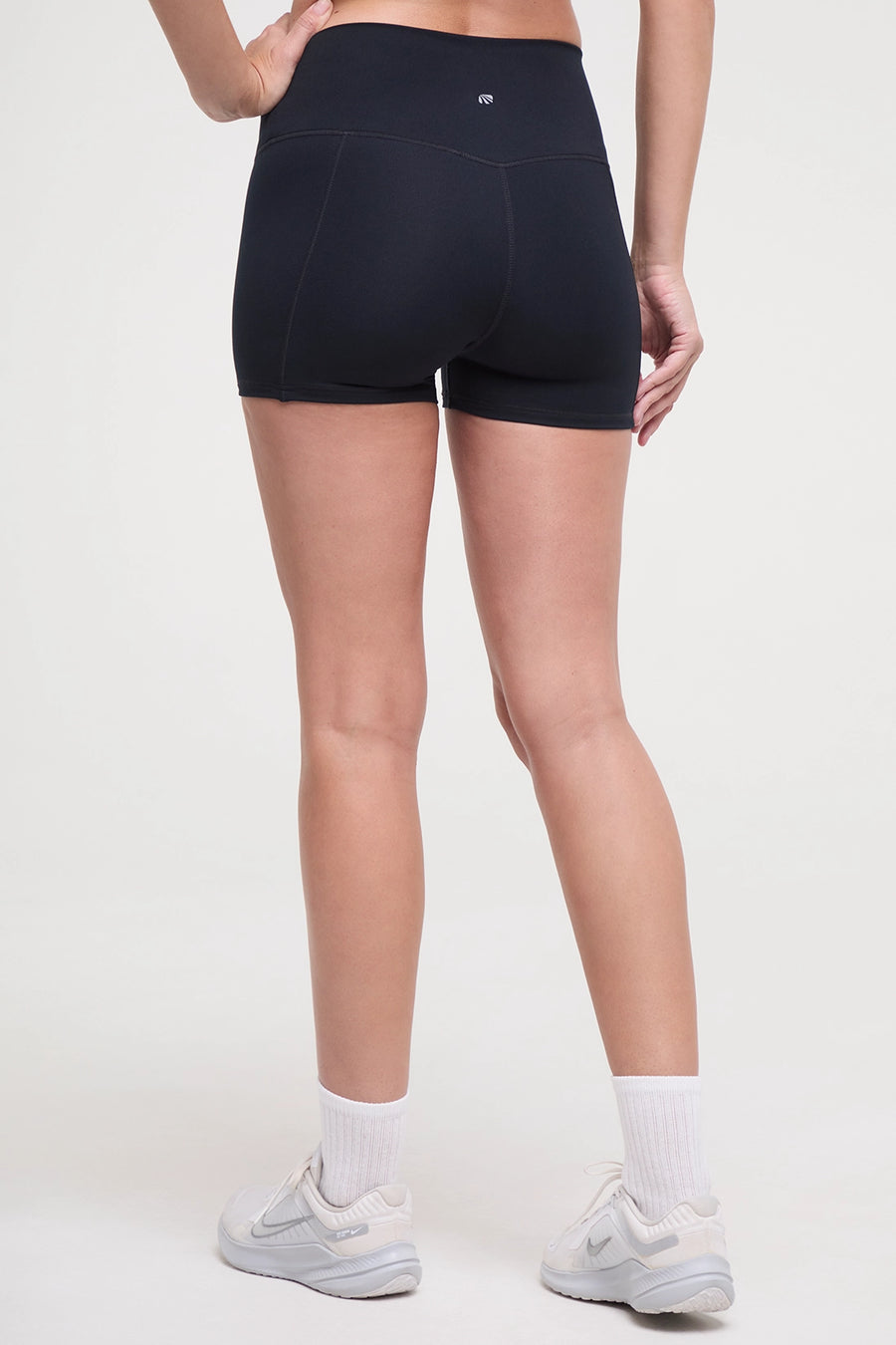 Back view of woman wearing black athletic shorts and white sneakers on a light background. #color_black