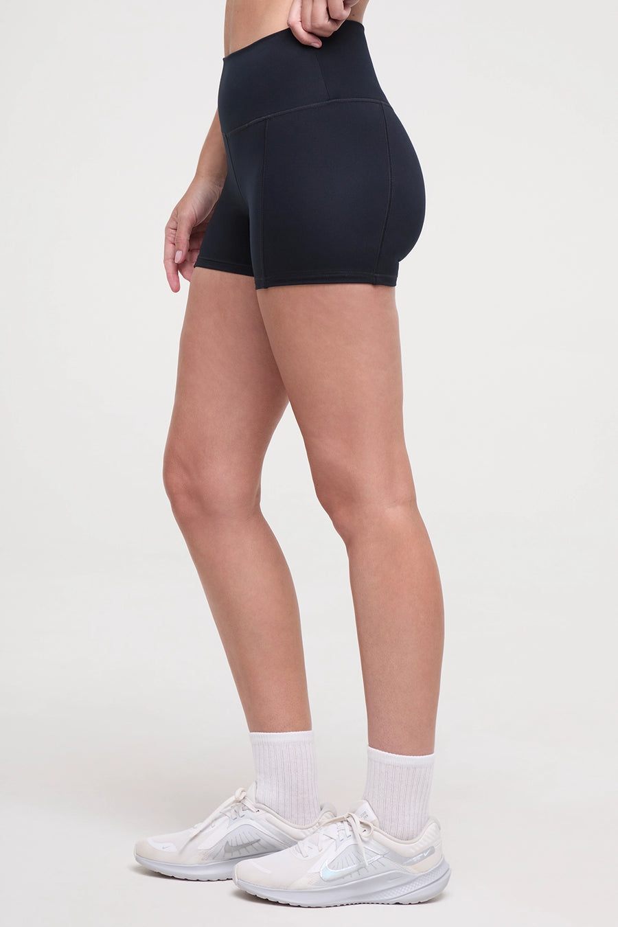 Side view of woman wearing black athletic shorts and white sneakers on a light background. #color_black