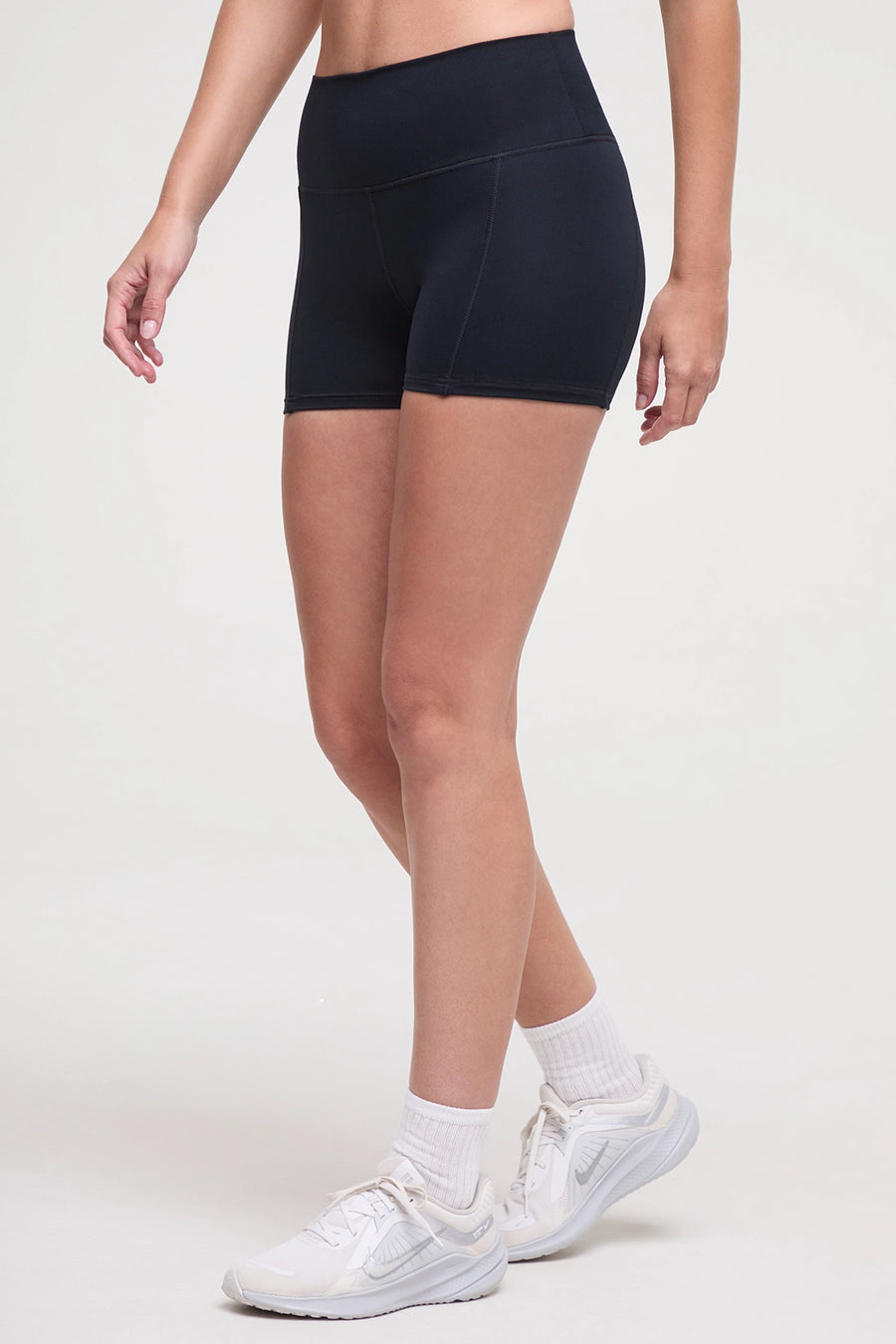 Front view of woman wearing black athletic shorts and white sneakers on a light background. #color_black