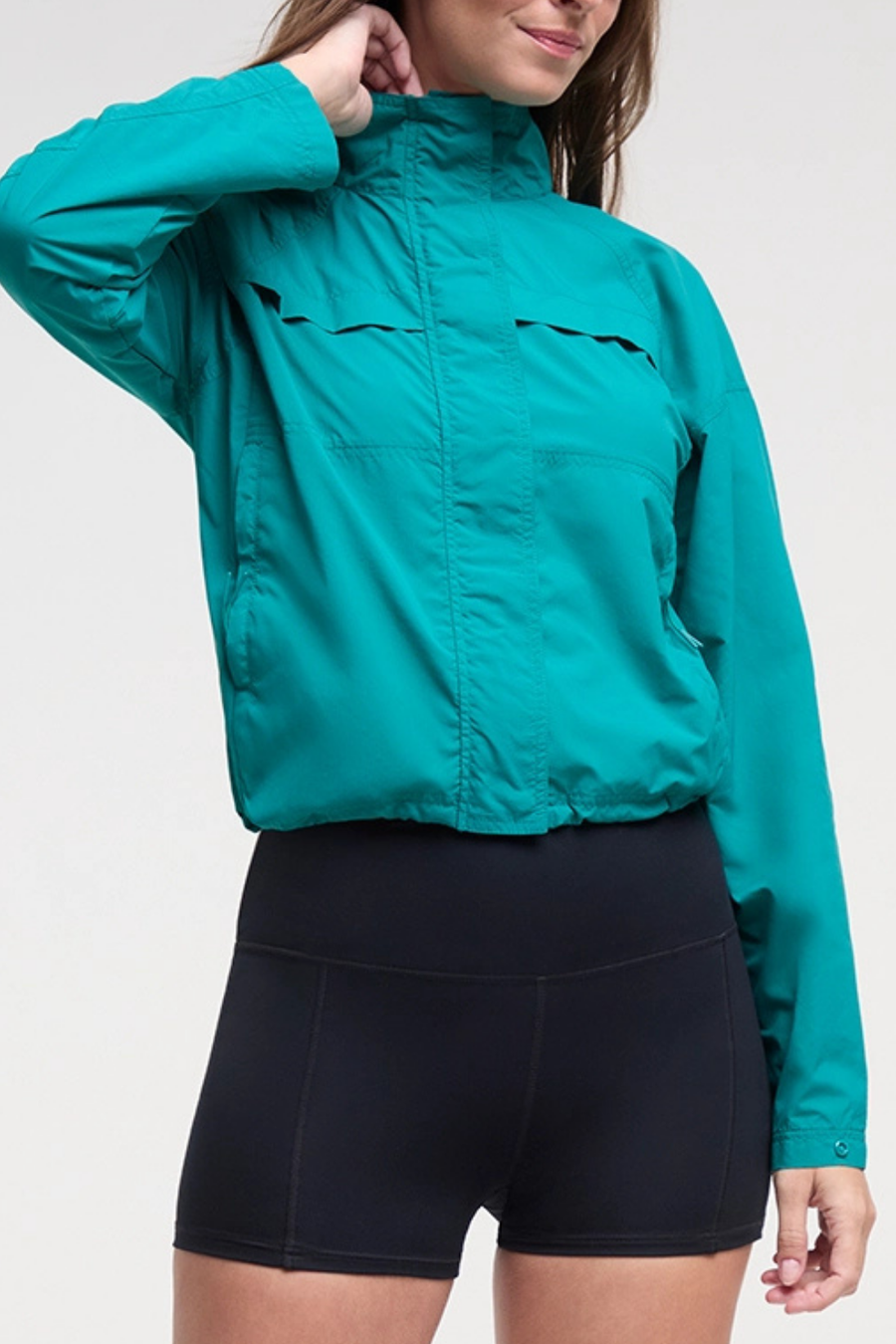 Front view of woman wearing a teal jacket and black shorts on a white background. #color_quetzal-green
