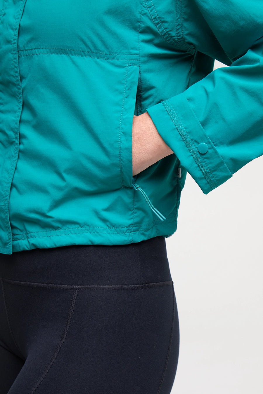 Close up view of woman wearing a teal jacket with zipper pocket and black shorts on a white background. #color_quetzal-green