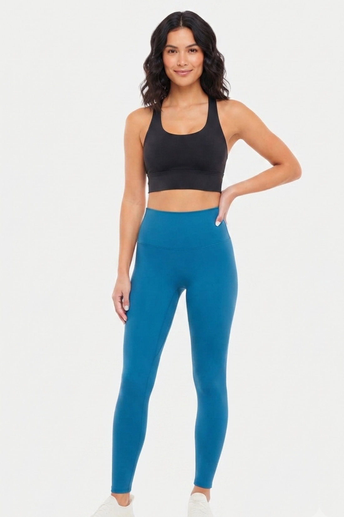 A woman modeling a black sports bra and teal blue high-waisted leggings against a light gray studio background. 