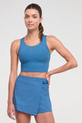 A woman modeling a matching athletic set consisting of a blue scoop-neck crop top and a blue wrap-style mini skirt, photographed against a light gray studio background. #color_midnight