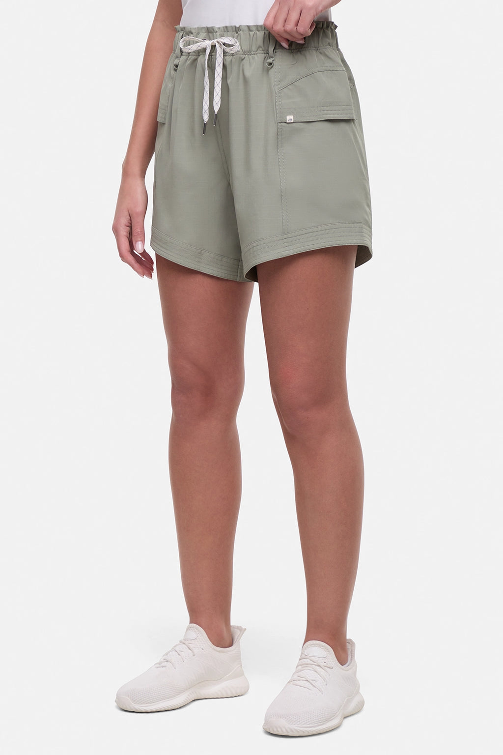 Side view of woman wearing sage green shorts with white drawstring waist and with side pockets and white sneakers on a white background. #color_shadow
