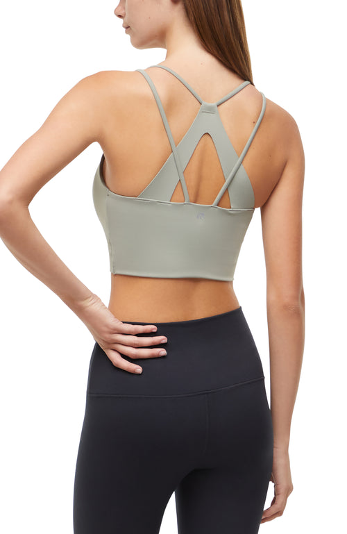 Back view of woman wearing a light sage green sports bra with criss-cross straps and black leggings on a white background. #color_shadow