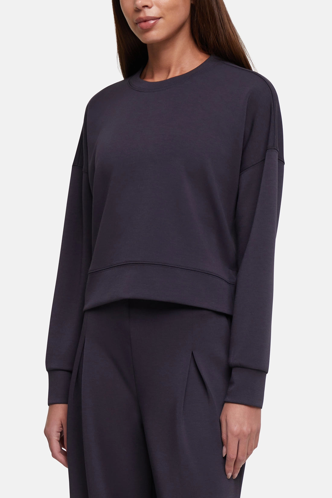 A woman wearing a dark charcoal gray cropped crewneck sweatshirt and matching sweatpants against a light gray studio background. 