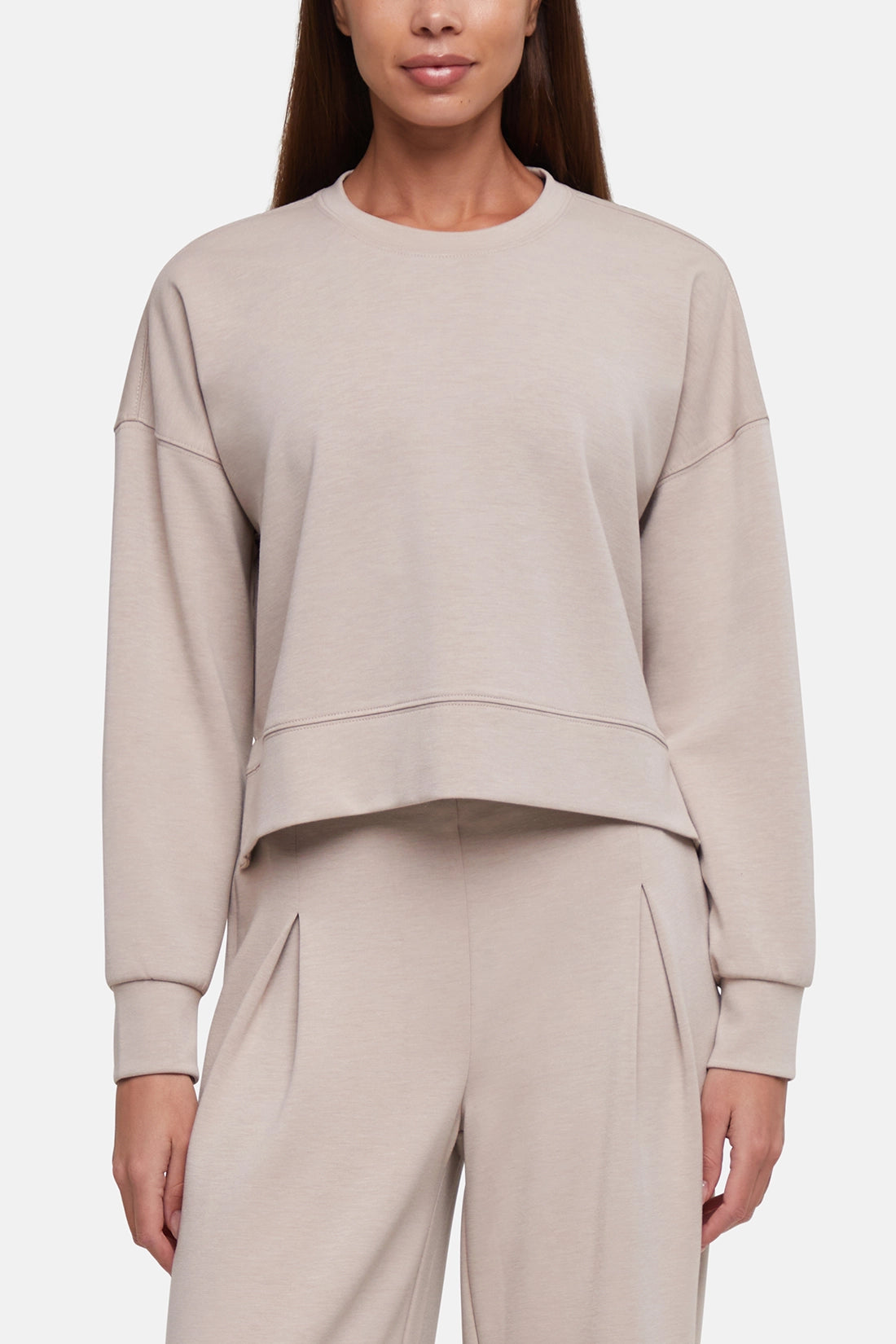 A woman wearing a light beige cropped crewneck sweatshirt and matching wide-leg sweatpants against a plain light gray studio background. 