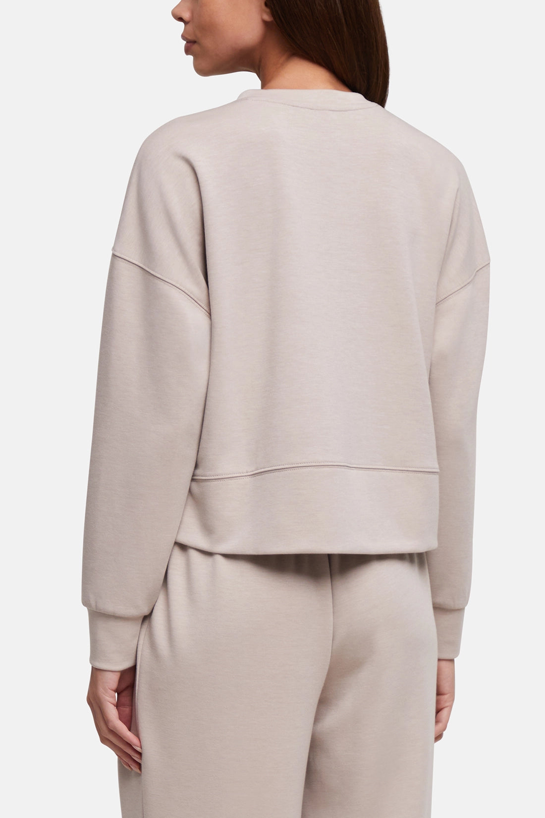 A woman shown from the back wearing a light beige cropped sweatshirt and matching sweatpants against a plain light gray studio background. 