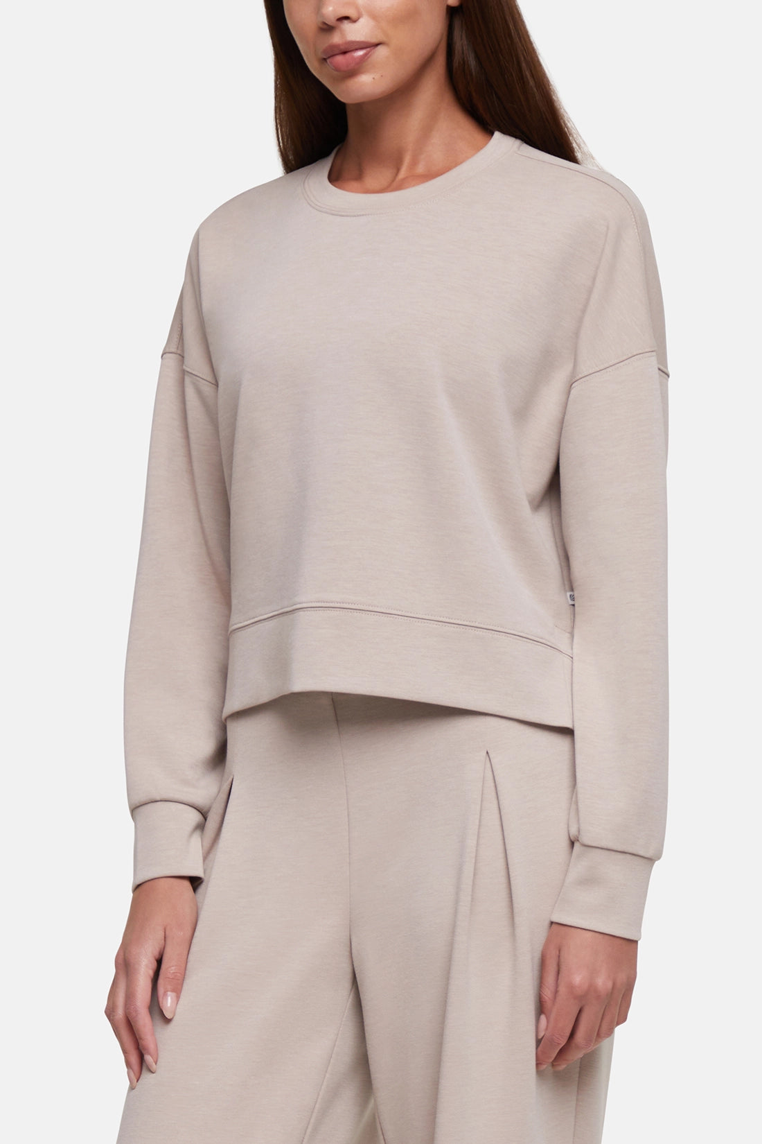 A woman wearing a light beige cropped crewneck sweatshirt and matching wide-leg sweatpants against a plain light gray studio background. 