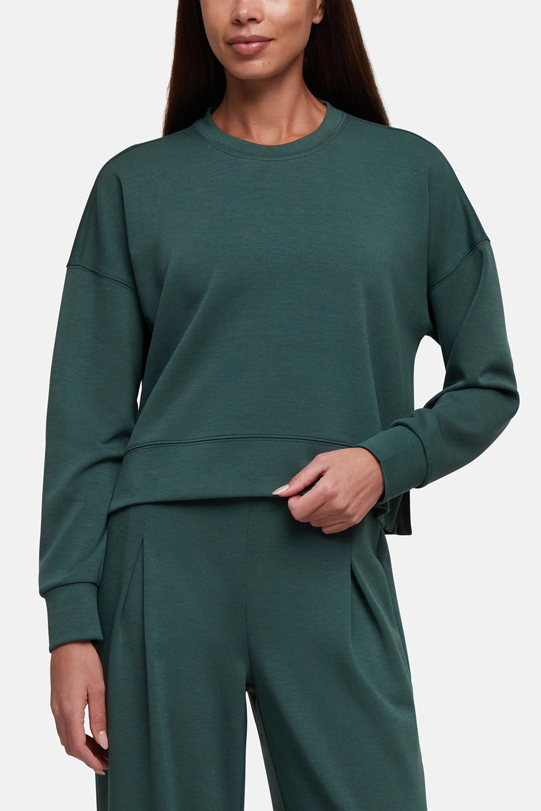 A woman wearing a dark green crewneck sweatshirt and matching wide-leg sweatpants against a light gray studio background. 