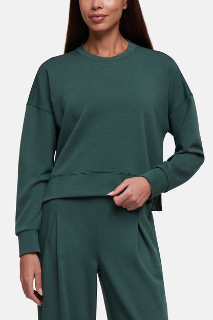 A woman wearing a dark green crewneck sweatshirt and matching wide-leg sweatpants against a light gray studio background. 