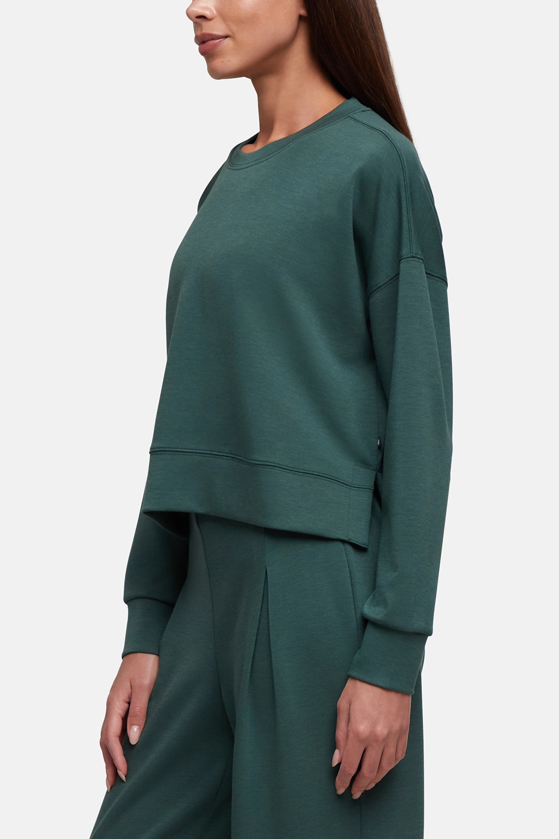 A woman wearing a dark green crewneck sweatshirt and matching wide-leg sweatpants, shown in a side-profile view against a light gray studio background. 
