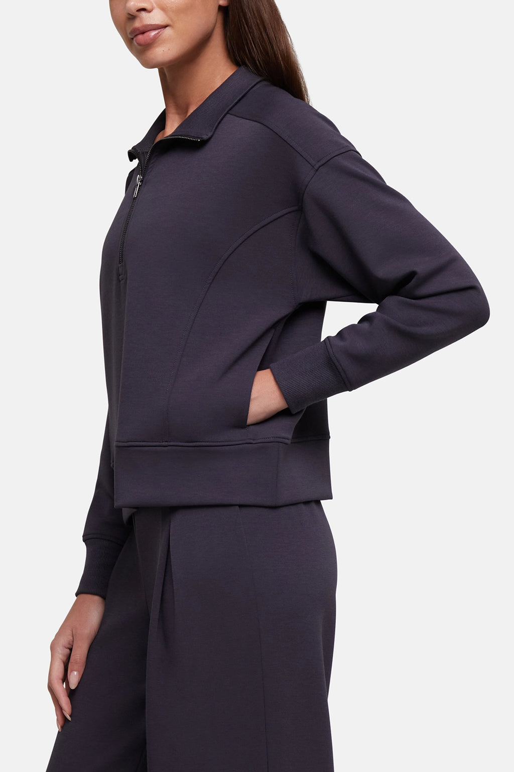 A woman wearing a dark charcoal gray zip-up sweatshirt with a half placket and matching dark gray sweatpants, shown from the side against a light gray studio background.