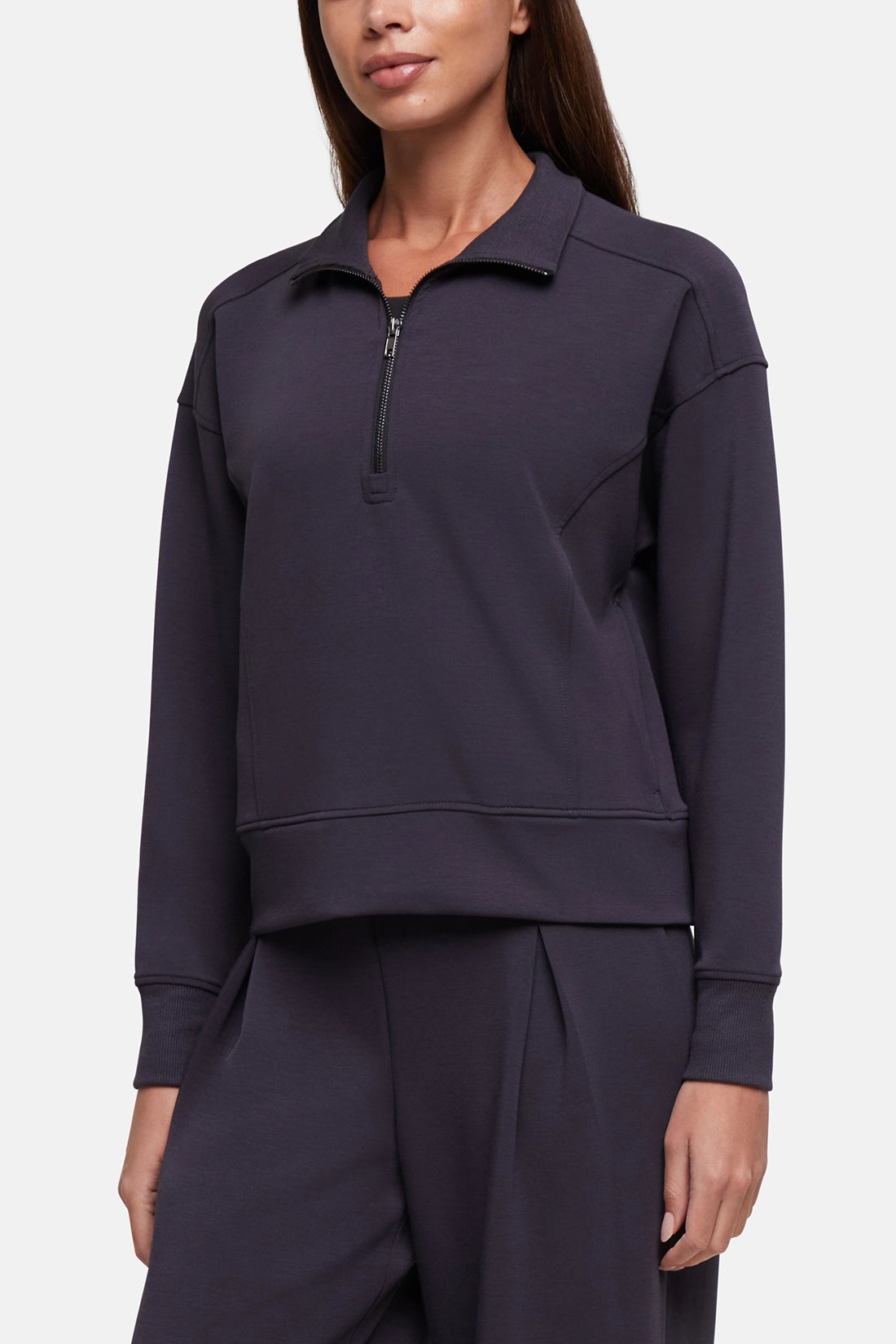 A woman wearing a dark purple quarter-zip sweatshirt and matching sweatpants against a light gray studio background.