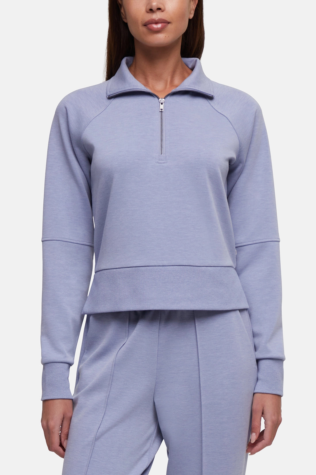 A woman wearing a light periwinkle blue half-zip pullover sweatshirt and matching sweatpants against a light gray studio background.  