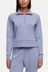 A woman wearing a light periwinkle blue half-zip pullover sweatshirt and matching sweatpants against a light gray studio background.  