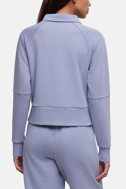 A model shown from the back wearing a light periwinkle blue cropped raglan sweatshirt and matching sweatpants against a plain light gray studio background. 