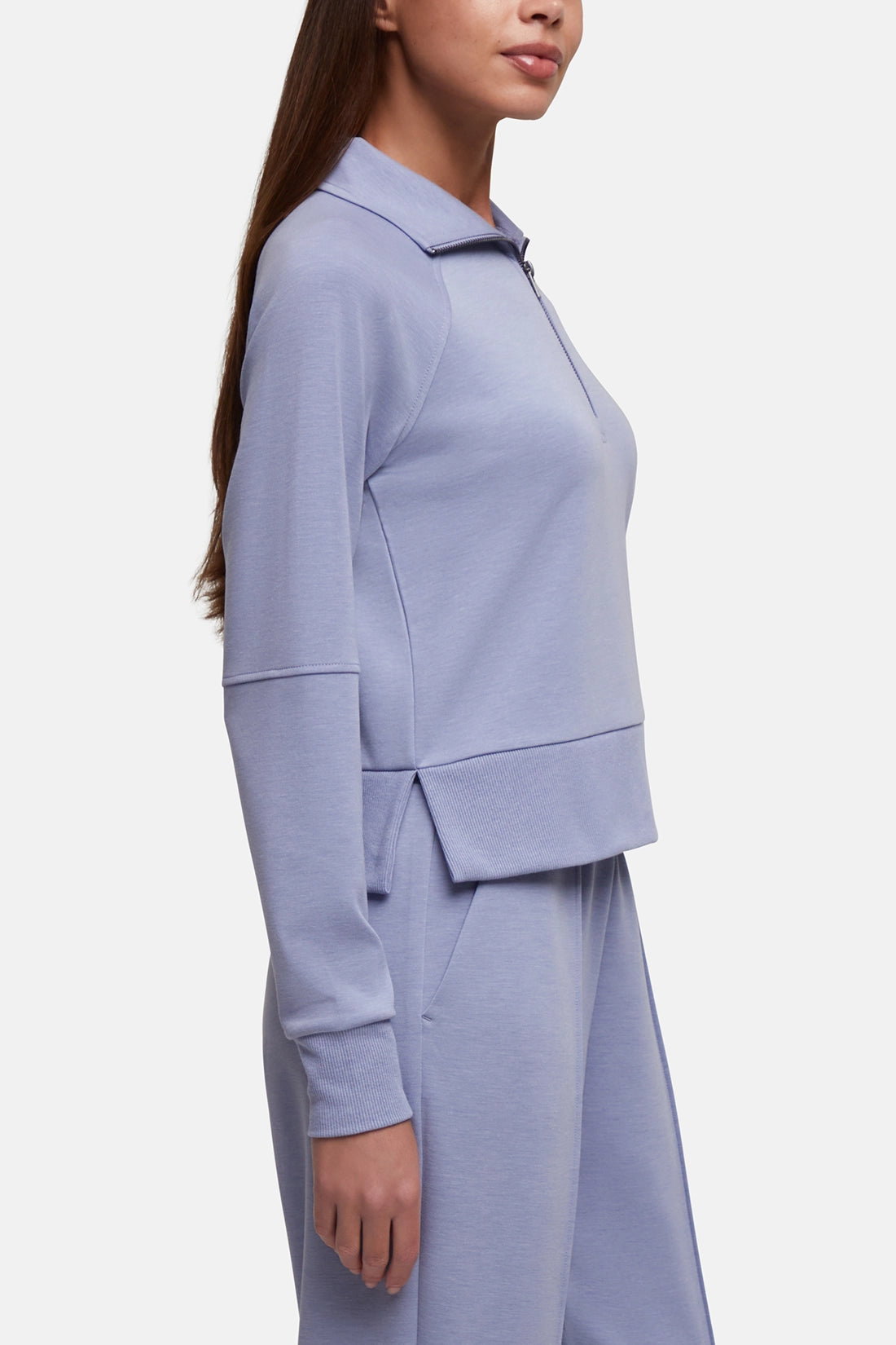 A woman shown in side profile wearing a light periwinkle blue matching knit set consisting of a cropped long-sleeve top with a mock neck and matching wide-leg trousers, against a plain light gray background. 
