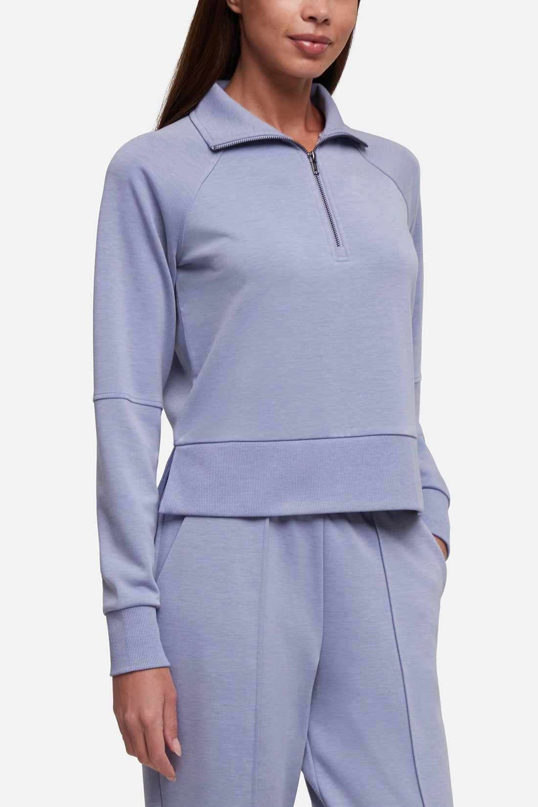 A woman wearing a light periwinkle blue matching tracksuit, featuring a long-sleeve quarter-zip pullover and matching jogger pants, against a plain light gray studio background. 