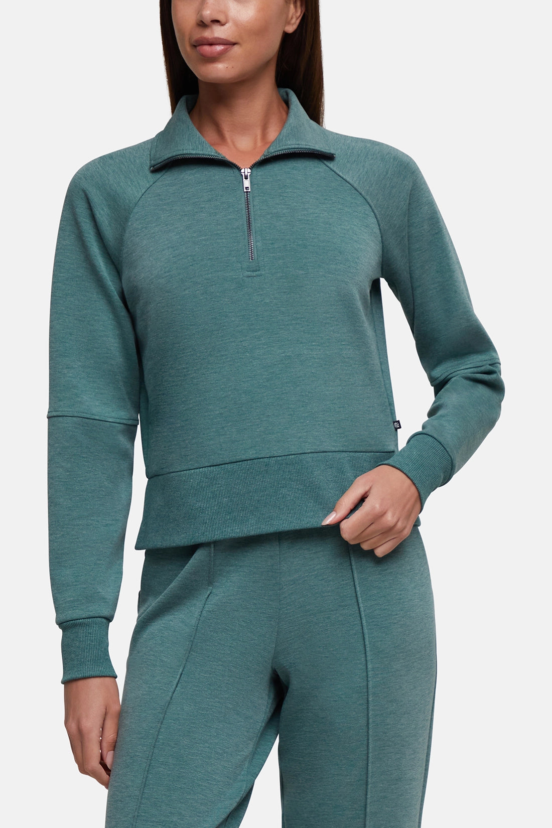 A woman wearing a matching teal green athletic set consisting of a cropped quarter-zip long-sleeve top and coordinating jogger pants, shown against a light gray studio background. 