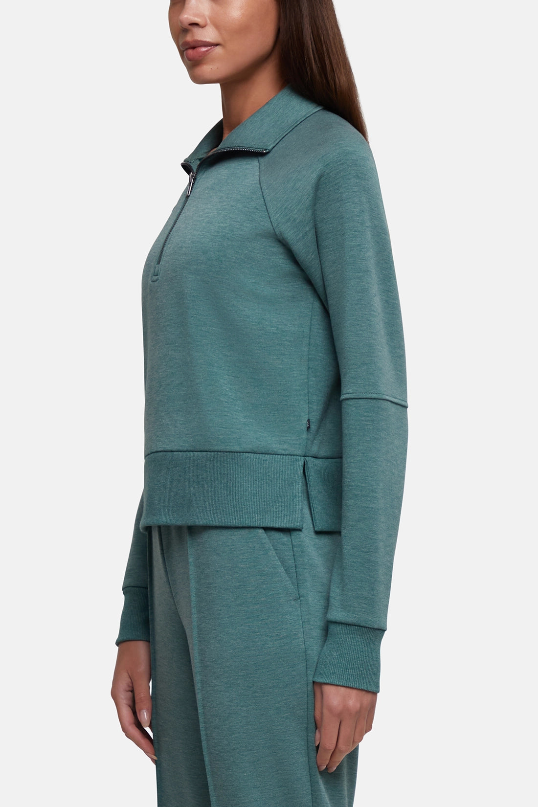 A woman wearing a matching teal green two-piece tracksuit, featuring a long-sleeve quarter-zip pullover top and coordinating pants, shown from a side profile against a light gray studio background. 
