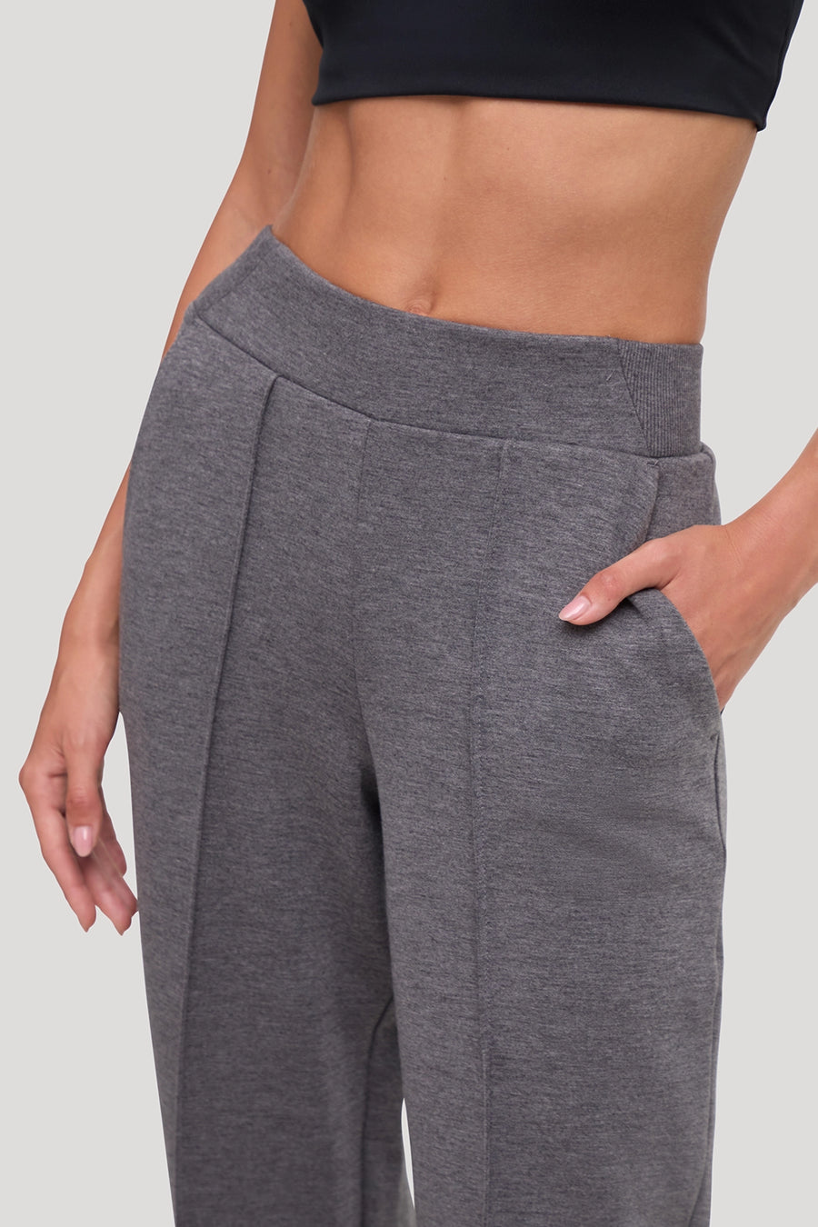 A close-up of a model wearing heather gray tapered-leg sweatpants with a pleated front and side pockets, paired with a black sports bra against a light gray studio background. #color_heather-black
