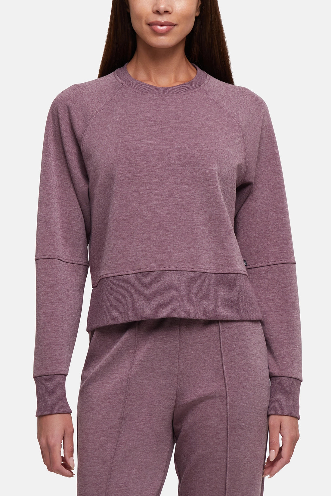 A woman wearing a heathered dusty mauve crewneck sweatshirt and matching sweatpants against a light gray studio background. #color_heather-fig