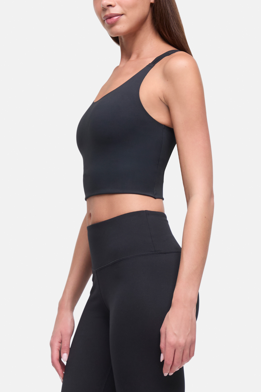 A woman wearing a black one-shoulder crop long bra and matching high-waisted leggings, shown from the side against a light gray studio background. #color_black