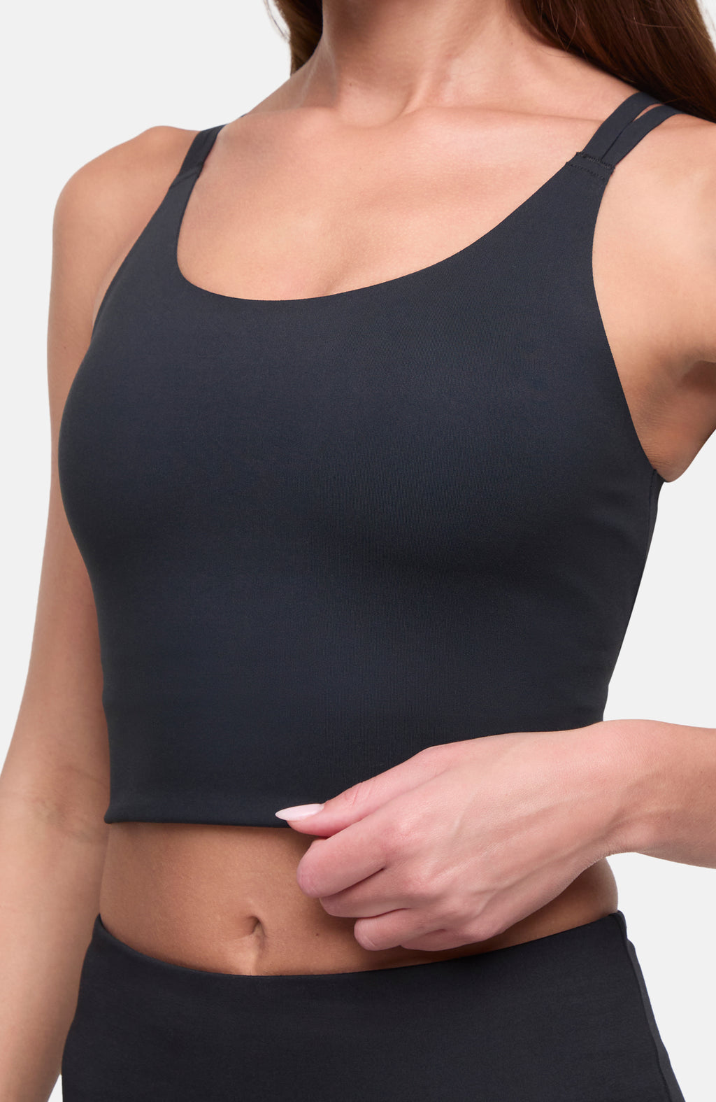 A close-up of a woman wearing a dark charcoal gray scoop-neck crop long bra and matching bottoms, with her hand lifting the hem of the top to show the fabric. #color_black