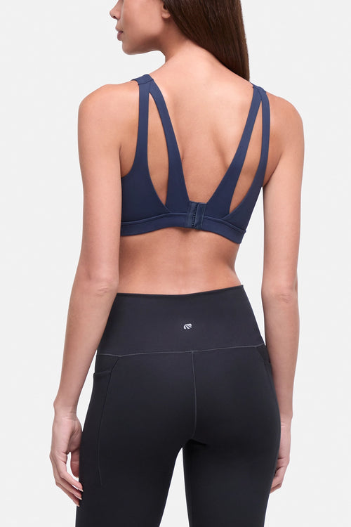 A woman shown from the back wearing a navy blue sports bra with cutout back straps and black high-waisted leggings against a light gray studio background. #color_total-eclipse