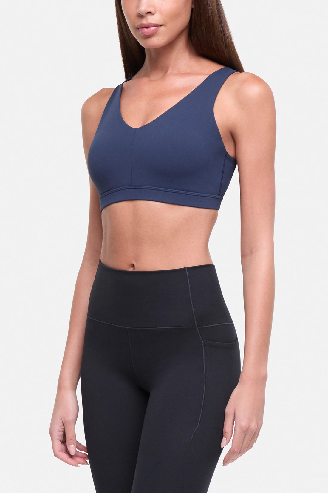 A woman modeling a navy blue sports bra and black high-waisted leggings against a light gray studio background. #color_total-eclipse