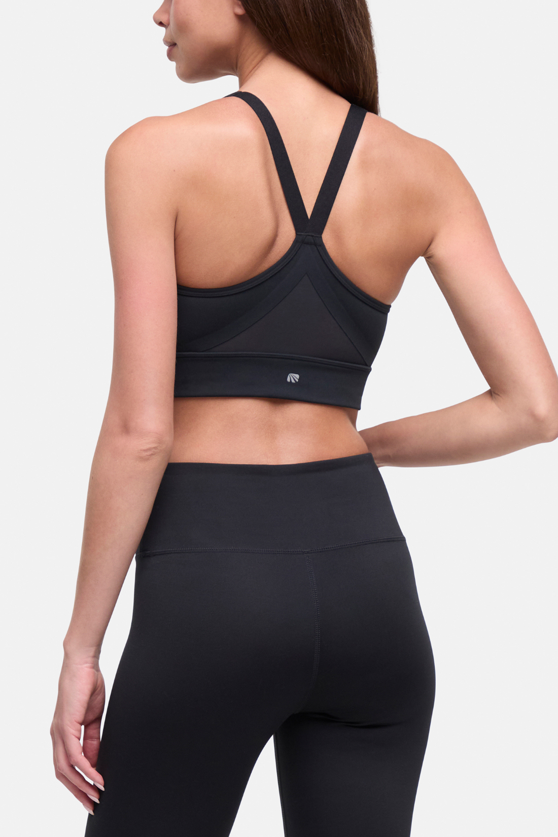Rear view of a woman wearing a black sports bra with crisscross back straps and black high-waisted leggings against a light gray studio background. #color_black