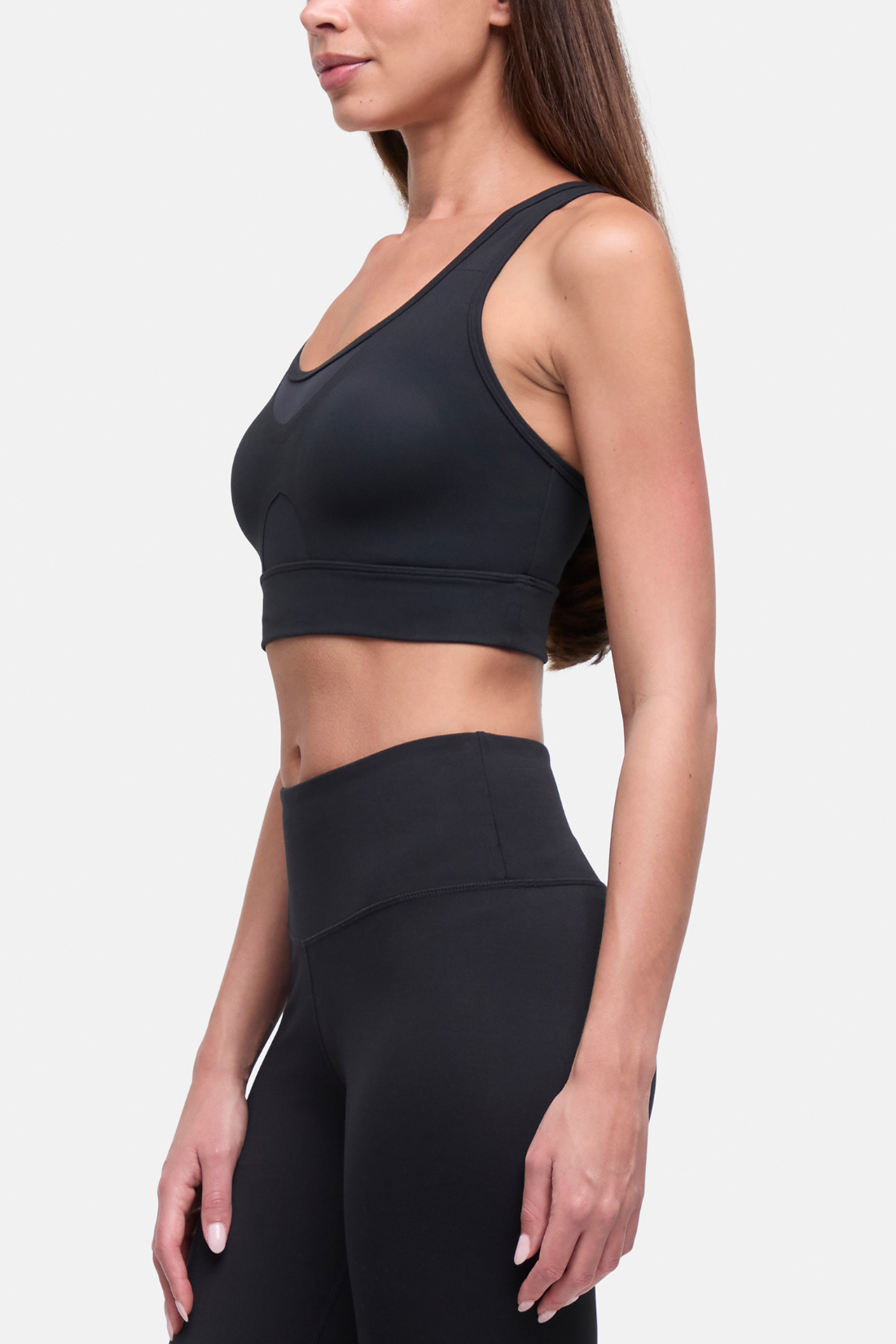 A woman wearing a black athletic sports bra and matching high-waisted leggings, shown from the side against a light gray studio background. #color_black