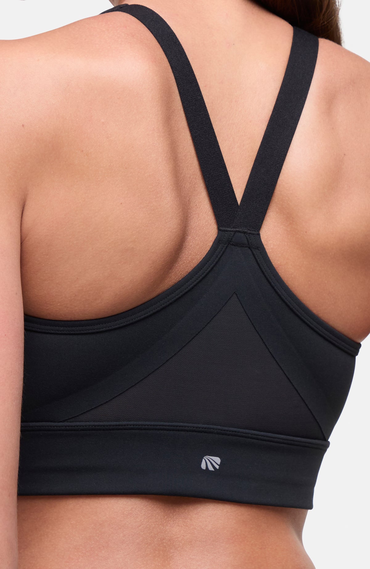 Close-up back view of a woman wearing a black sports bra with wide underarm panels and crossover straps, shown against a light neutral background. #color_black