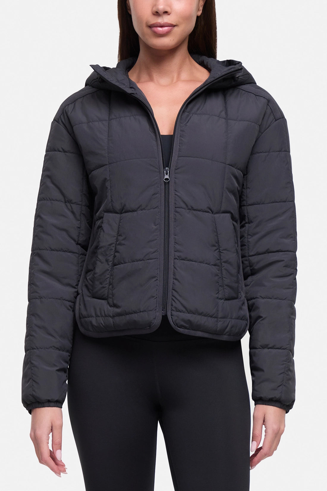 A woman wearing a dark gray hooded puffer jacket with a front zipper and quilted stitching, paired with black leggings, against a light gray studio background.