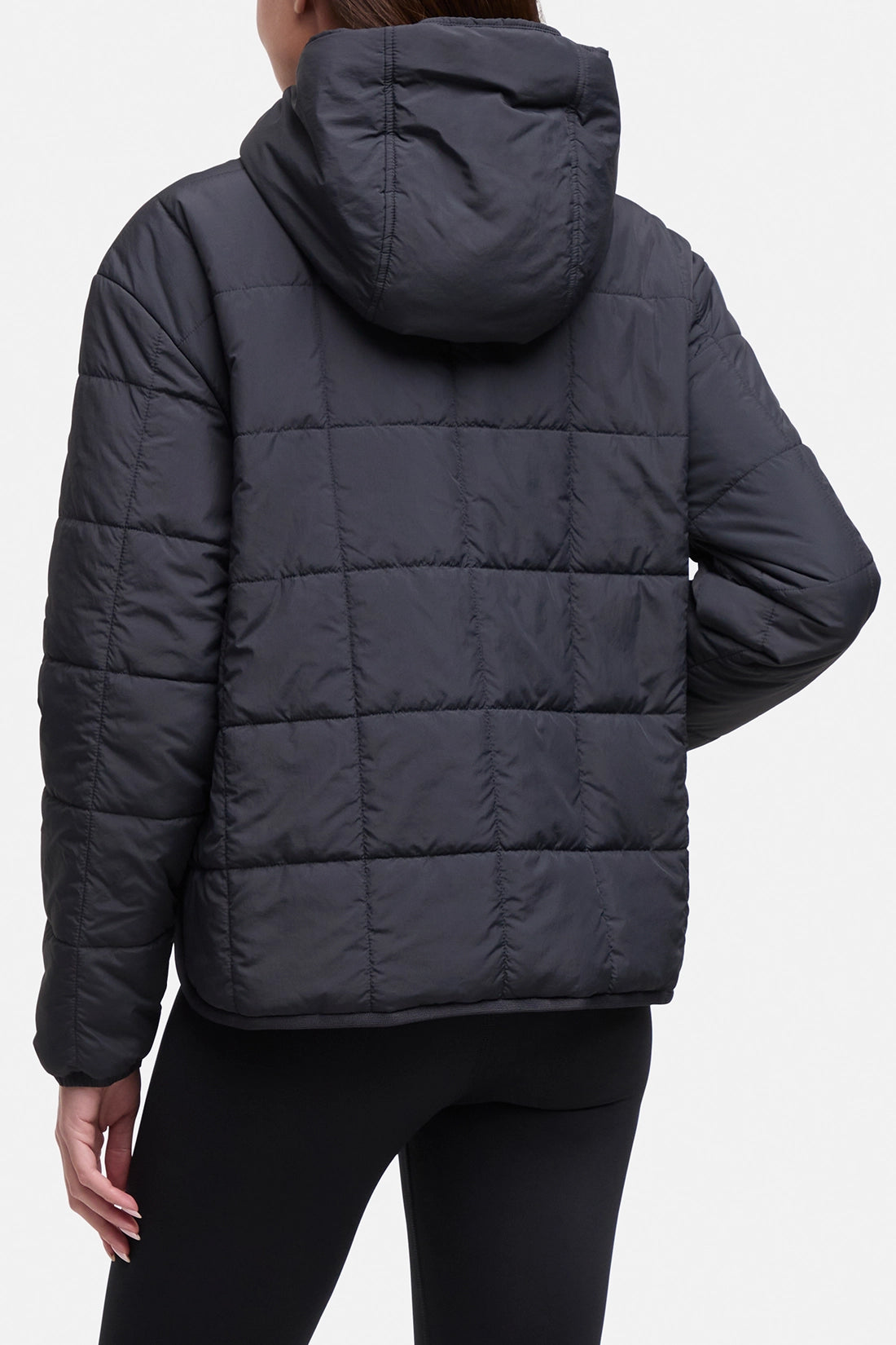 A person shown from the back wearing a dark charcoal gray quilted puffer jacket with a hood and black leggings against a light gray studio background.