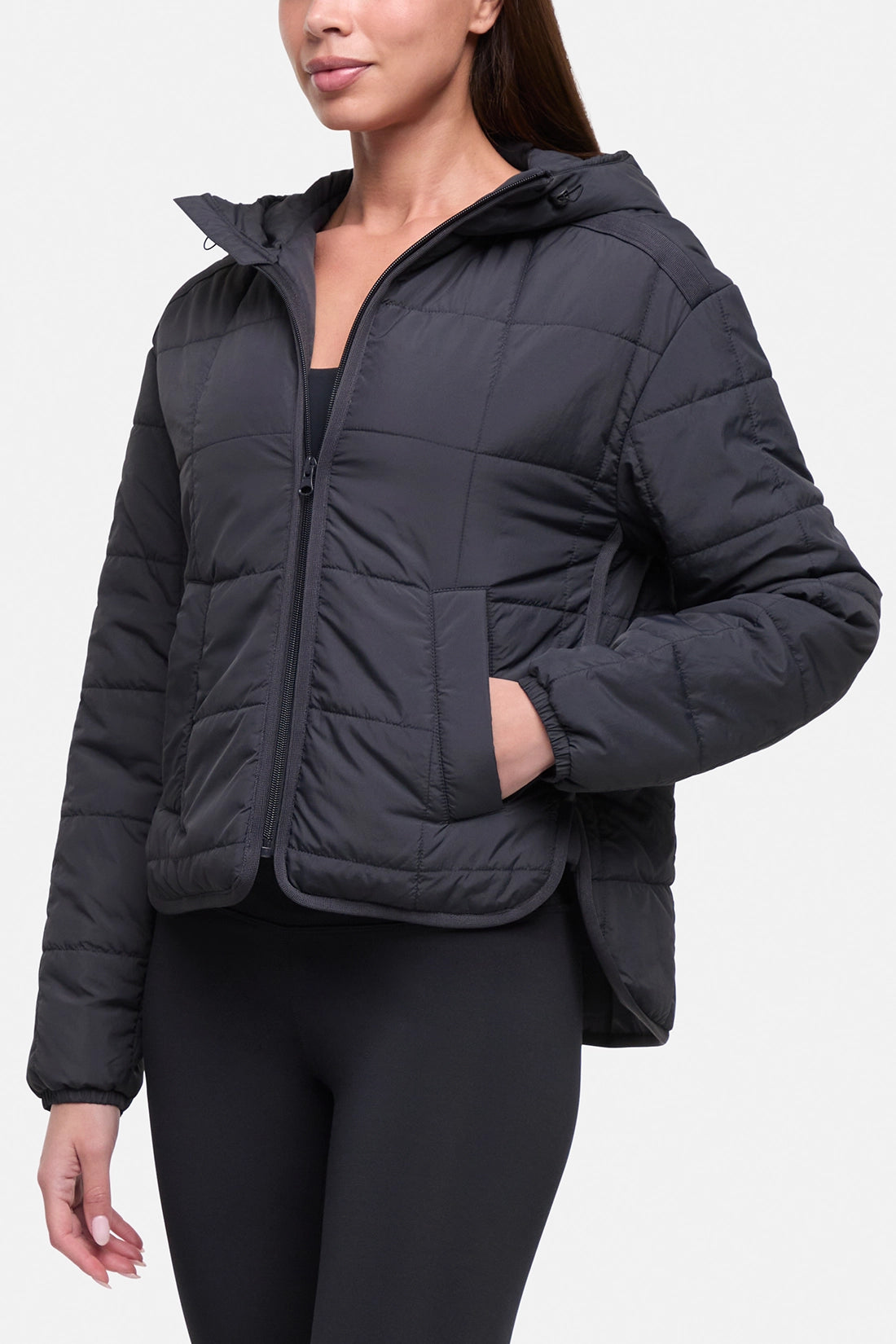 A woman wearing a dark gray quilted puffer jacket with a hood and front zipper, paired with black leggings, shown against a light gray studio background.