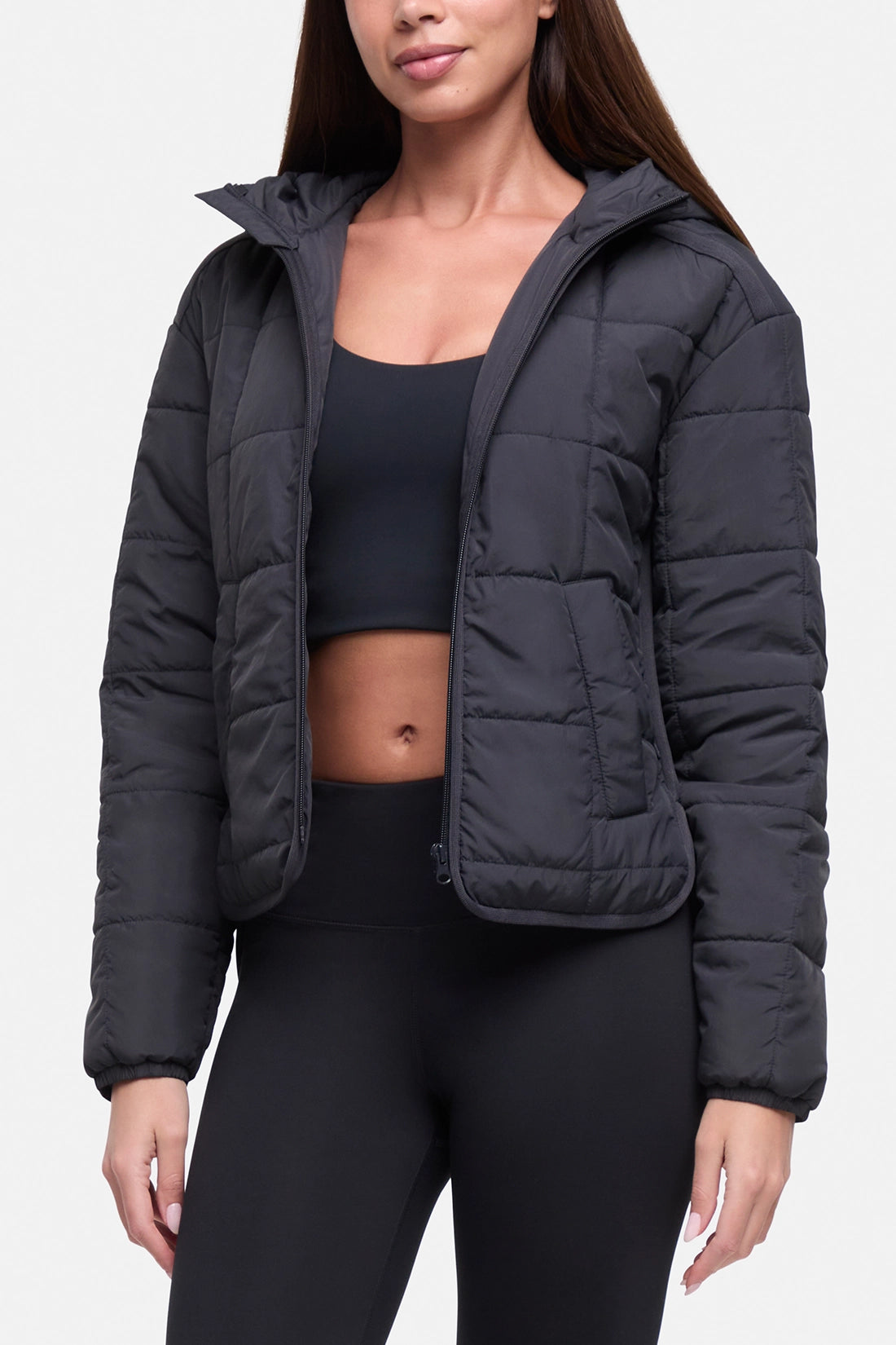 A woman wearing a black cropped puffer jacket with a high collar, styled over a black sports bra and black leggings, against a light gray studio background.