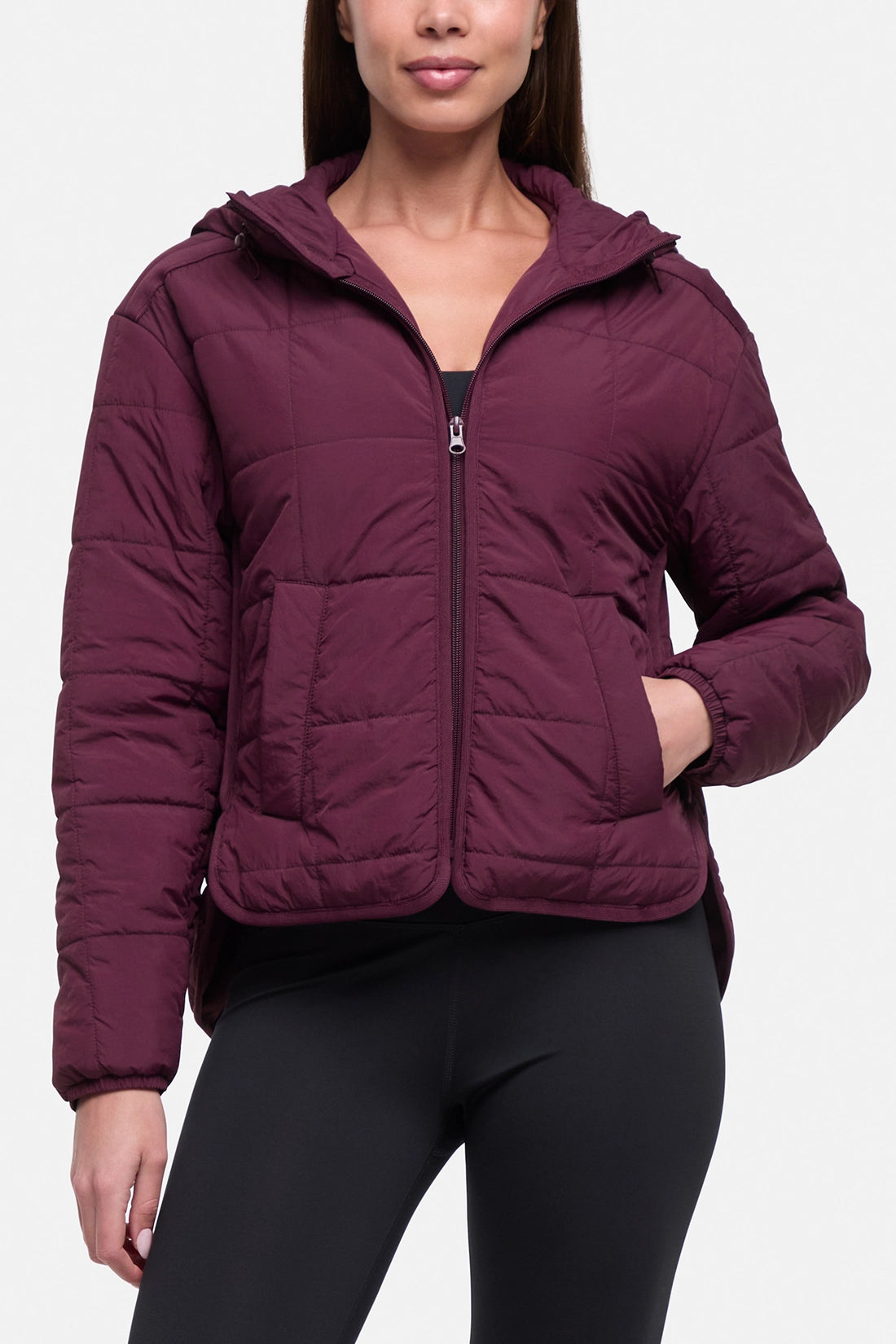 A woman wearing a cropped, quilted puffer jacket in deep burgundy with a front zipper, hood, and zippered pockets, paired with black leggings, against a light gray studio background. #color_winetasting