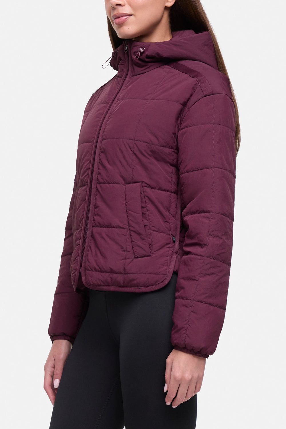 A woman wearing a cropped, quilted puffer jacket in a deep plum purple color, paired with black leggings, shown from a side profile against a light gray studio background.