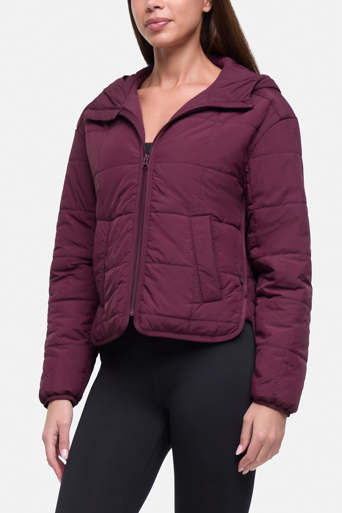 A woman wearing a cropped, quilted puffer jacket in deep burgundy with a front zipper and hood, paired with black leggings, against a plain light gray background.
