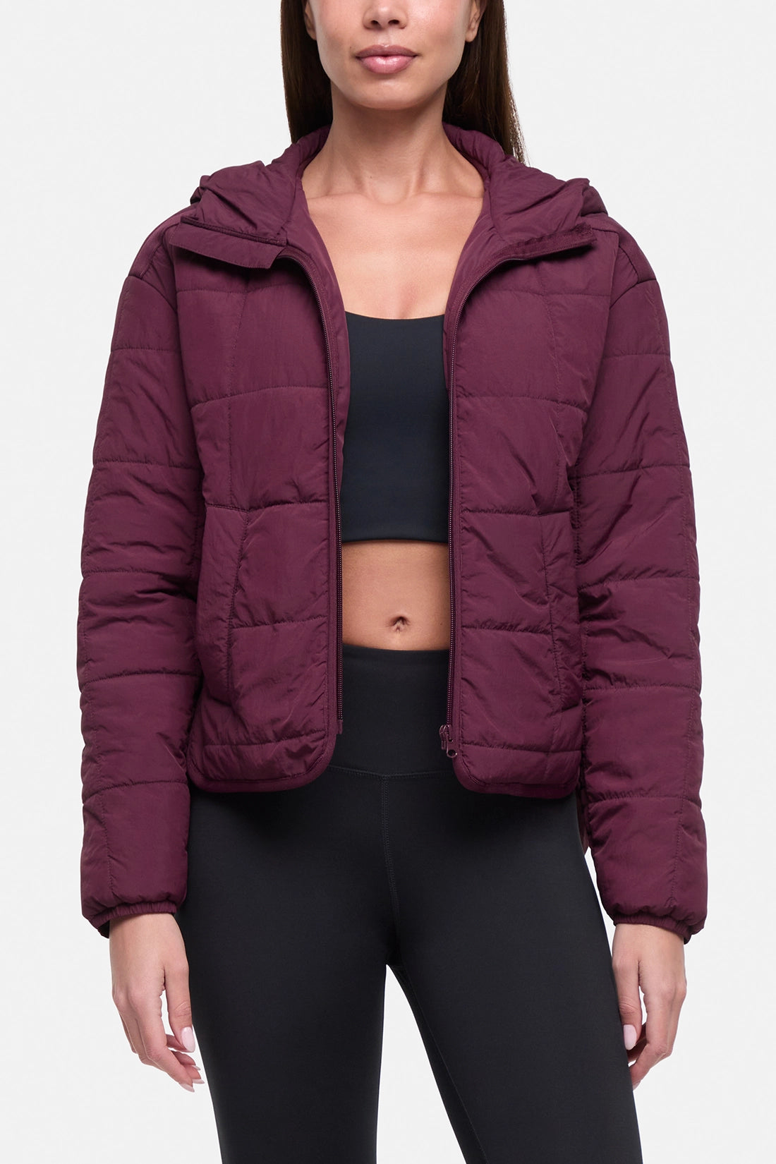 A woman wearing a cropped, quilted puffer jacket in a deep plum or burgundy shade, styled with a black sports bra and black leggings against a light gray studio background.
